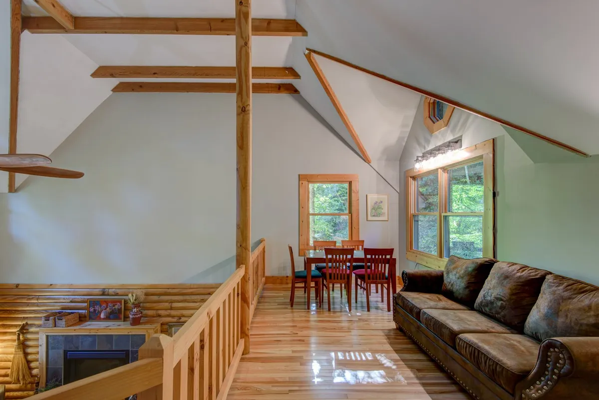 A sunlit interior features sturdy wooden beams, a plush sofa, a cozy dining area, and expansive windows. The atmosphere radiates warmth and comfort, inviting guests to gather for family meals, relax with a book, or simply bask in the golden light. The room’s open layout and natural materials evoke the homestead's signature welcoming feel.
