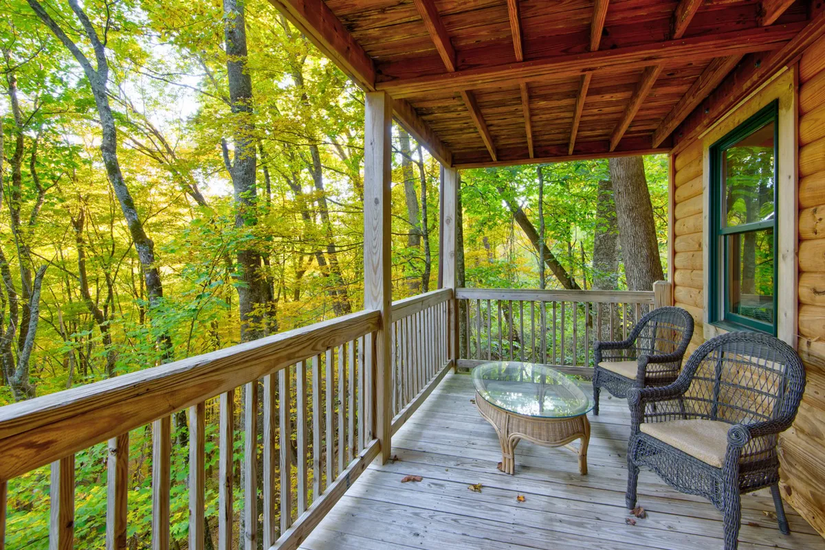 A serene wooden deck nestled among trees, furnished with a glass table and woven chairs for outdoor relaxation. The surrounding woodland and natural tranquility are visible, offering a seamless extension of the indoor communal space.