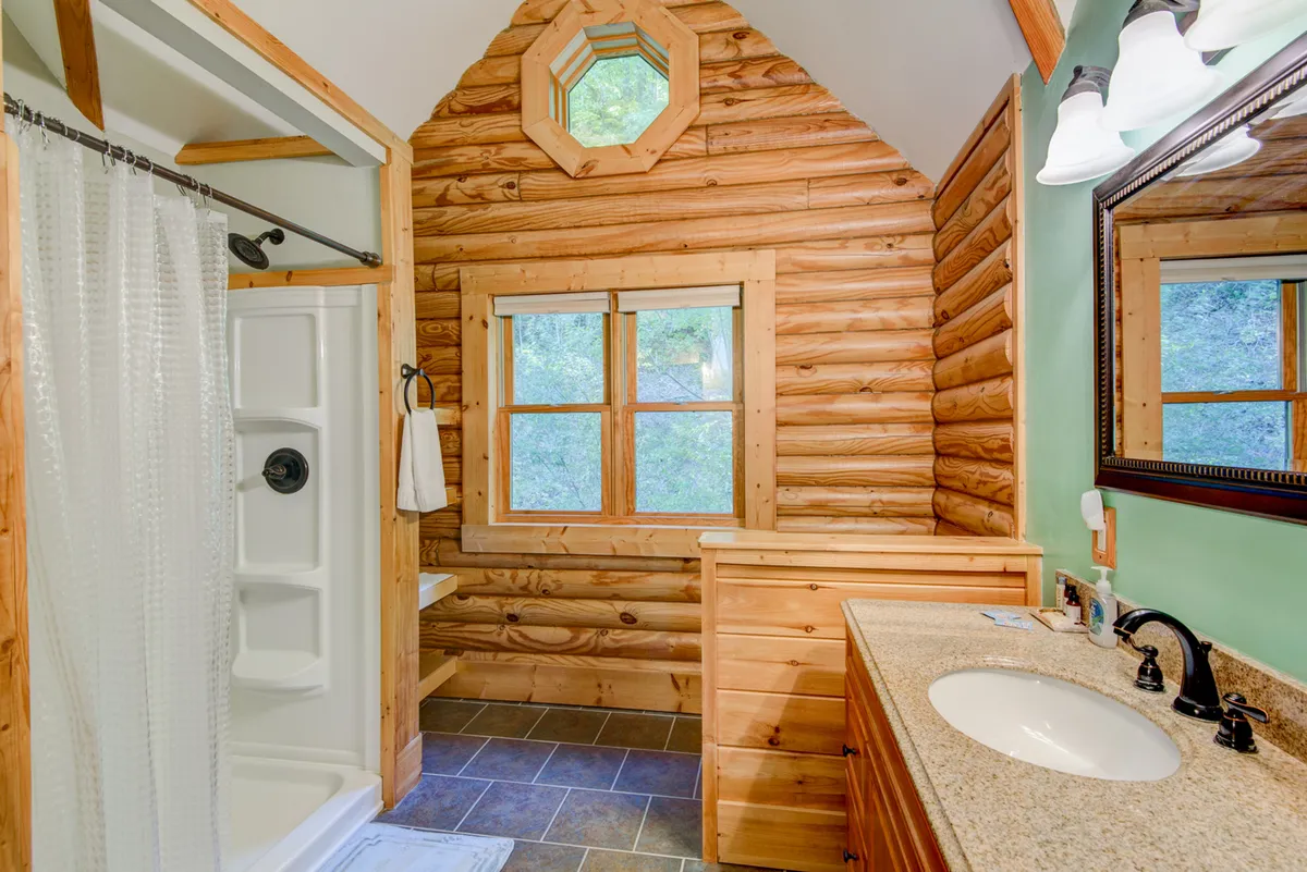 A tranquil bathroom with log walls, an octagonal window bringing in sunlight, a spacious vanity with sink, and a shower area. The natural wood and light create a spa-like experience.