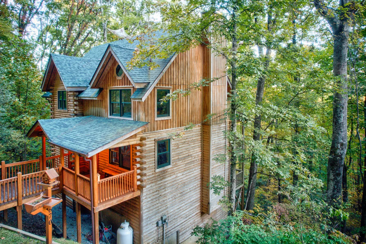 A broad view of the log cabin structure nestled among trees, with a visible balcony area and ample surrounding forest. The image captures both the house’s rustic charm and its direct access to outdoor adventure, with natural light bringing out the warmth of the wood.