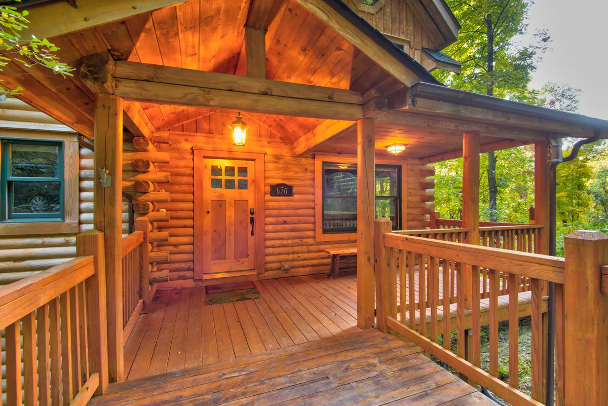A wide view of the welcoming log cabin entrance with a wooden railing, front door, and a cozy porch area. Warm lighting and natural wood textures create a sense of arrival and hospitality, inviting guests into the mountain retreat.