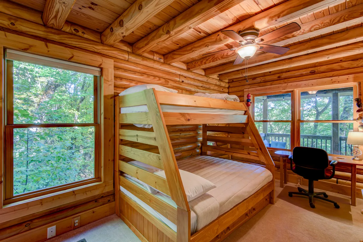 A warm, welcoming bedroom with a twin-over-full bunk bed, large windows, wooden ceiling, and a small desk with a chair. The room is filled with natural light and features rustic wood elements, making it ideal for children or small families.