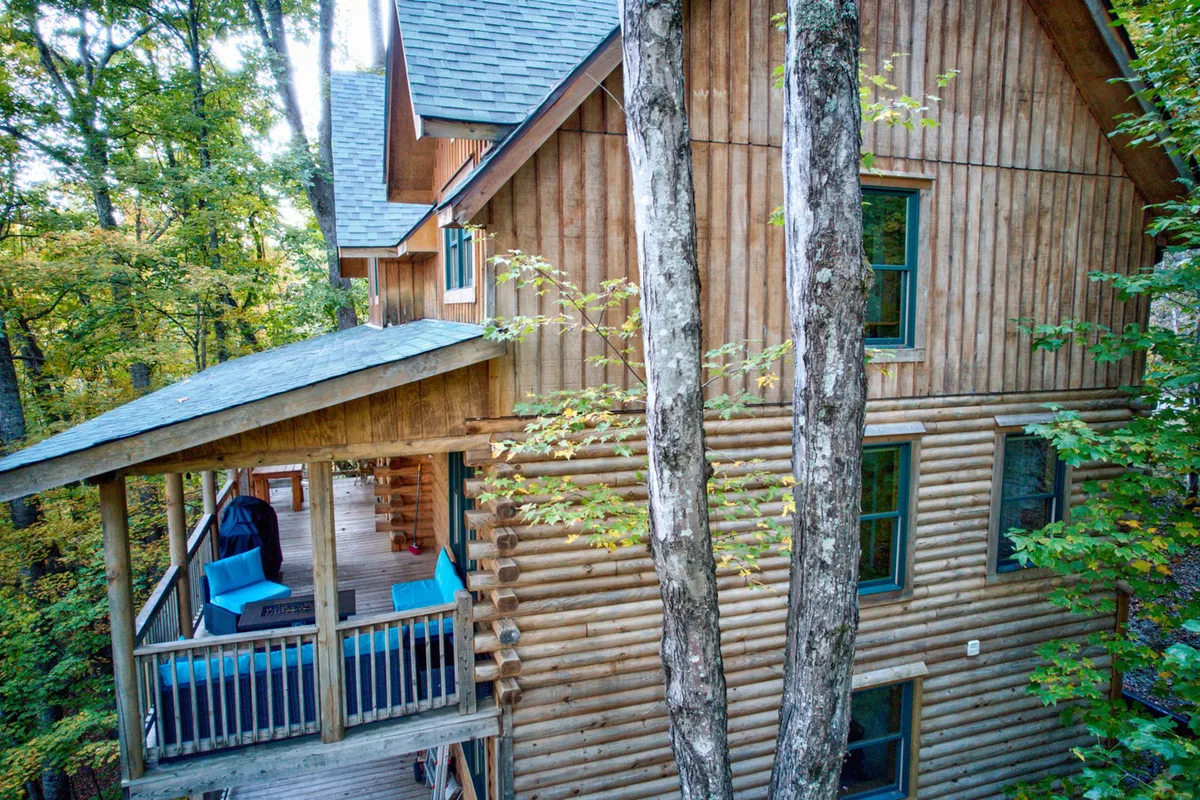 The image captures the cabin's wooden log siding, a cozy blue outdoor seating arrangement, and surrounding trees. The inviting porch setting, nestled among greenery, is perfect for relaxed mornings or social evenings, emphasizing the continuity of comfort and special moments available to loyal guests.