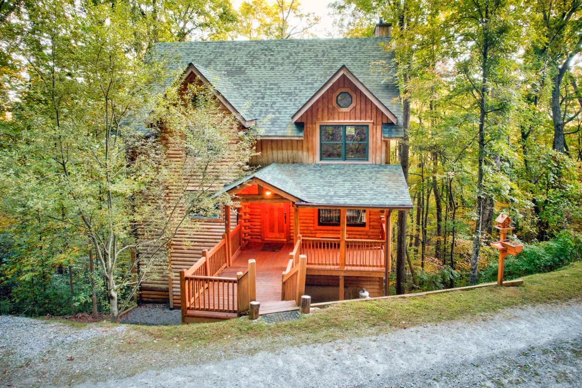 This exterior image captures the log cabin structure of Hideaway Homestead, complete with a welcoming wooden deck and a front porch nestled among tall trees. The setting suggests an inviting, nature-immersed environment ideal for family gatherings and outdoor relaxation, echoing the property's proximity to local attractions.