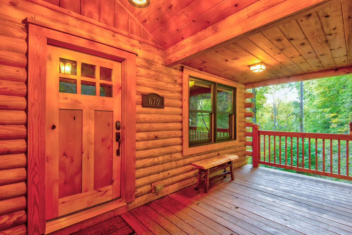 The log cabin's inviting wooden front door and bench sit under a covered porch, surrounded by pine trees. The seating area hints at quiet moments spent enjoying the fresh mountain air or gearing up for a walk.