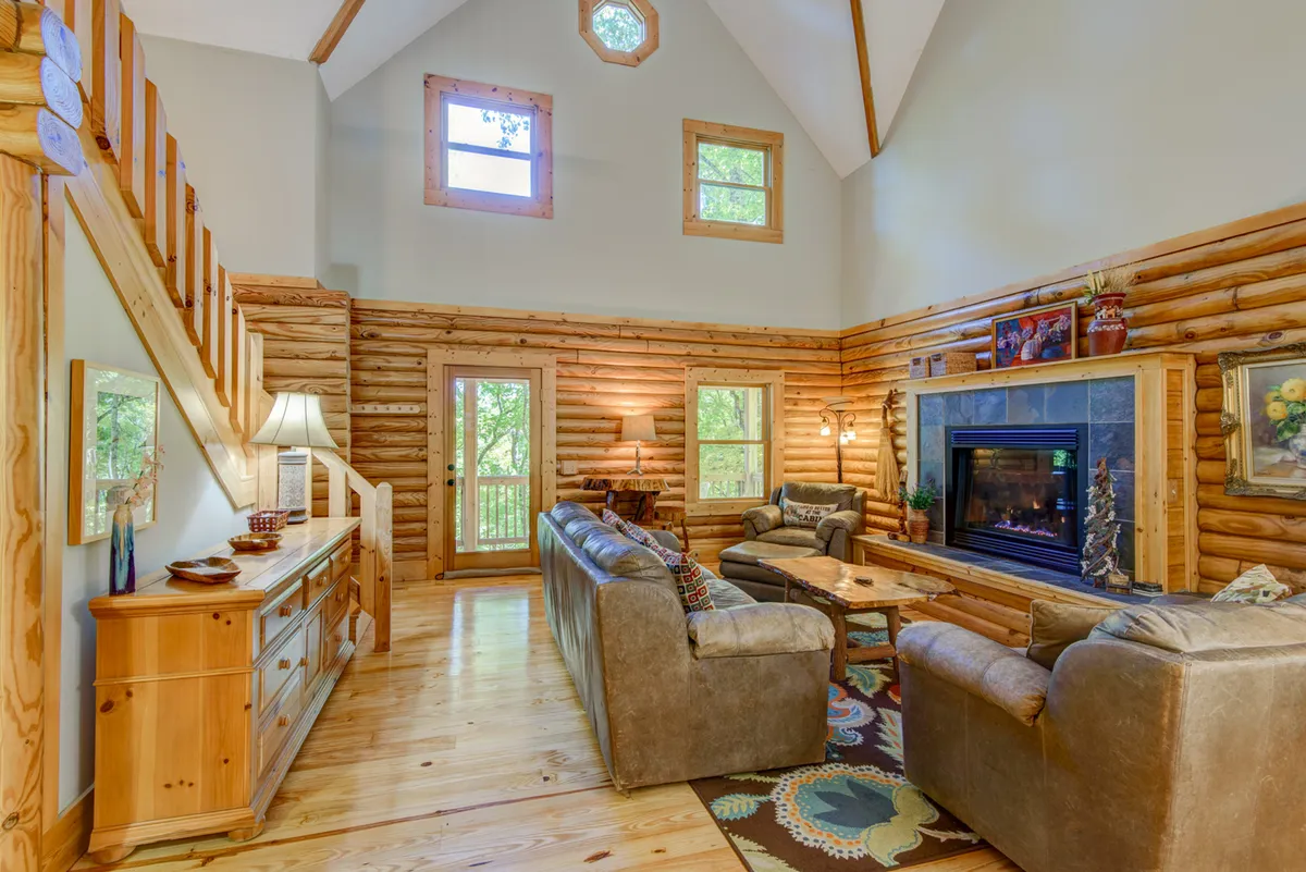 A warm, inviting living room with a fireplace, tall windows looking out to the woods, a wooden staircase, and comfortable sofas. The space feels perfect for cozying up after an outdoor adventure, with ample room for pets and family.