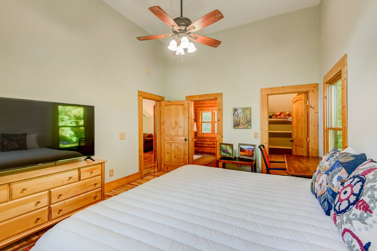 A cozy bedroom featuring a bed with white bedding and decorative pillows, a wooden dresser with a flatscreen TV atop, and wooden doors leading to adjacent rooms. Natural light filters in, enhancing the inviting and restful atmosphere.